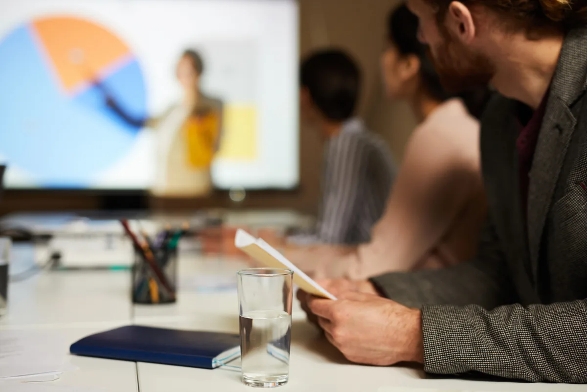 Sala riunioni: una relatrice illustra un grafico proiettato mentre i partecipanti seguono dal tavolo