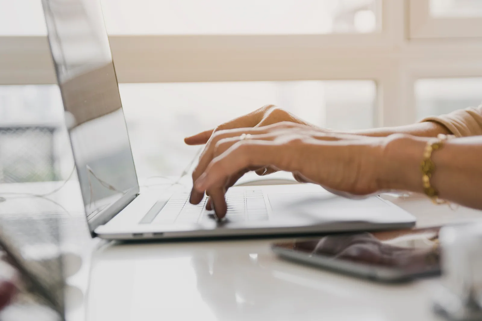 Mani che digitano sulla tastiera di un computer portatile, accanto a uno smartphone su una scrivania chiara