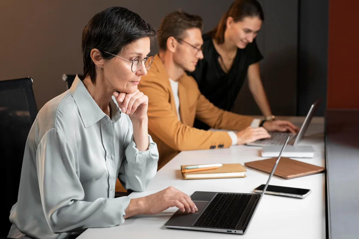 Donna con gli occhiali al computer portatile, seduta a un tavolo insieme a due colleghi